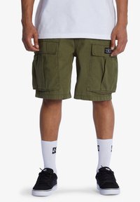 Olivegrüne Cargo-Shorts aus einem strukturierten Stoff mit großen Seitentaschen. Kombiniert mit schwarzen Sneakers und weißen Socken mit Logo.