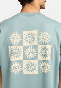 Hellesblaues Baumwoll-T-Shirt mit einem retro Sonnenmuster in Creme, das auf der Rückseite ein Rasterlayout mit neun Designs zeigt. Kurze Ärmel.