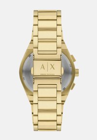 Kuldtooni roostevaba terasest kell, millel on lingitud rihm, ümmargune korpus ja graveeritud logo "A|X" taga.