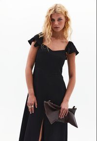 Vestido negro con tirantes de hombro con volantes, corsé ajustado y abertura lateral. La modelo sostiene un bolso marrón plegado, mostrando líneas elegantes y textura.