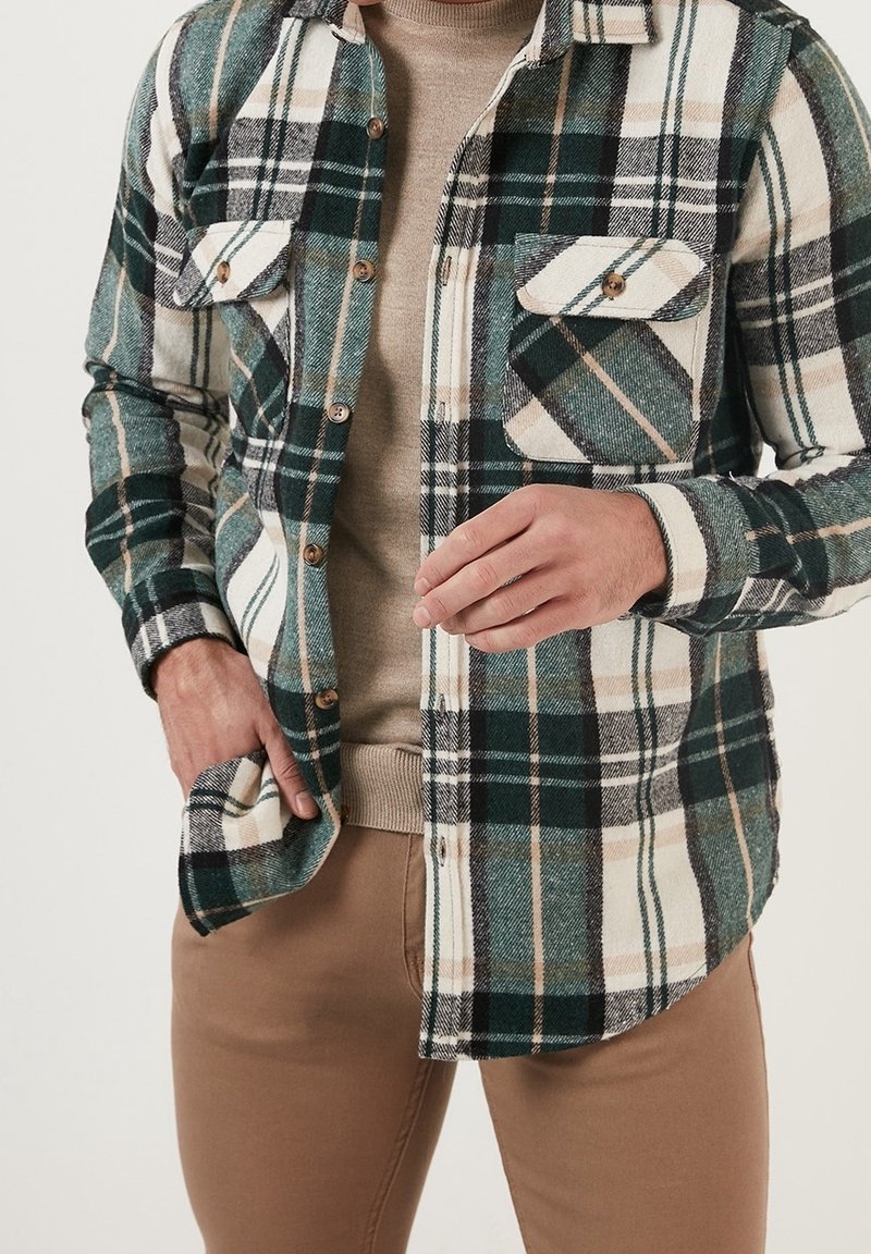 Kariertes Flanellhemd in Grün, Schwarz und Creme mit zwei Brusttaschen, durchgehender Knopfleiste und weicher Textur. Über einem beigen Top getragen.