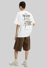 T-shirt bianca in cotone con una stampa grafica colorata sul retro, abbinata a pantaloni corti marroni e sneakers di colore chiaro.