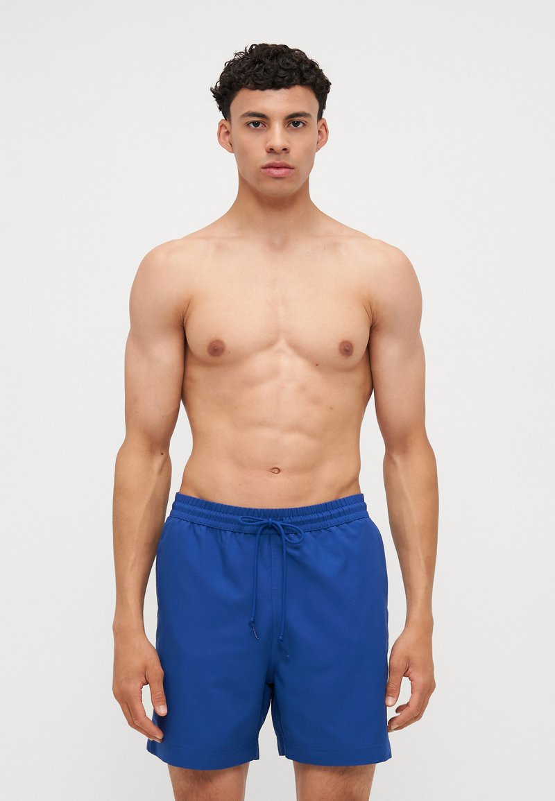 Blå svømmeshorts med elastisk talje og snøre. Lavet af glat materiale, med lige snit og sidesømme uden lommer.