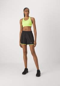 Sujetador deportivo en verde neón con un patrón triangular, combinado con pantalones cortos negros con detalles en neón y cinturilla elástica, llevados con zapatillas negras.