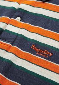 Koszulka polo w niebieskie, pomarańczowe, białe i zielone paski. Posiada kołnierzyk i zapięcie na guziki z przodu. Haftowane logo "SuperDry Athletics" w kolorze pomarańczowym.