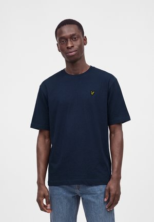 Giovane uomo con capelli corti che indossa una maglietta blu navy con un piccolo logo giallo a forma di uccello e jeans azzurri su sfondo bianco.
