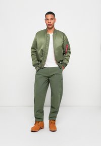 Olive green bomber jacket with zip pockets, white t-shirt, olive cargo pants, and brown lace-up boots. Smooth texture, casual fit.