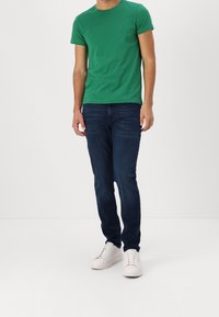 Grünes Baumwoll-T-Shirt mit kurzen Ärmeln, kombiniert mit dunkelblauen Jeans. Weiße Sneaker runden das Outfit ab und verleihen einen lässigen Look.