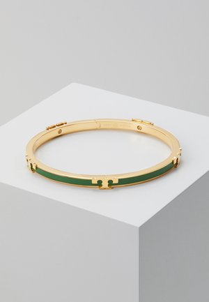 Goldener Armreif mit grünem Emaille-Einsatz und graviertem Verschluss, präsentiert auf einem weißen Podest vor neutralem Hintergrund.