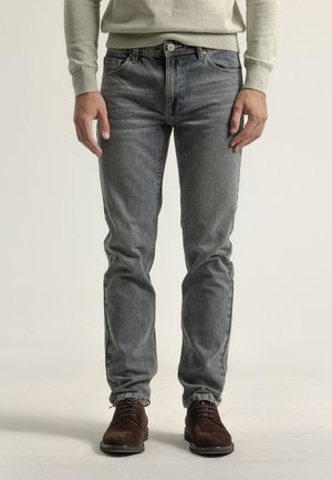 Jeans ajustados de color azul en denim desgastado con un diseño clásico de cinco bolsillos. Combinados con zapatos de ante marrón, mostrando un look informal.