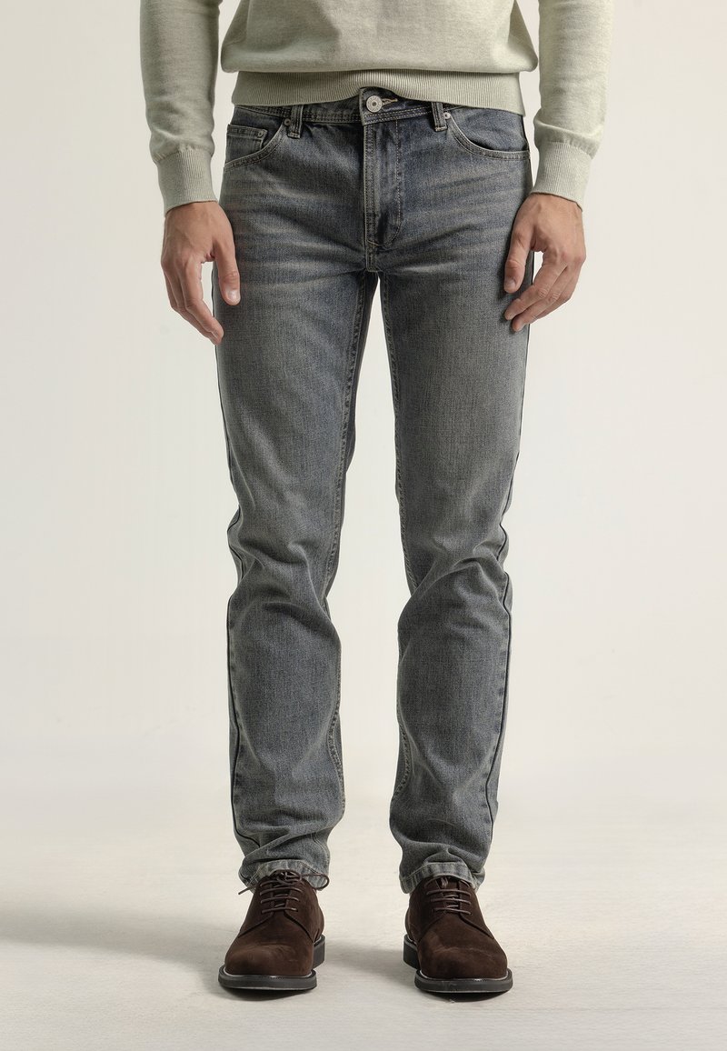 Jeans ajustados de color azul en denim desgastado con un diseño clásico de cinco bolsillos. Combinados con zapatos de ante marrón, mostrando un look informal.