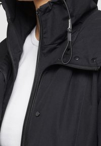 Musta hupullinen takki, jossa on vetoketju ja napit, päällä valkoinen ribbineuloksinen pyöreäkauluksinen paita, lähikuva ylävartalosta ja kaulasta.