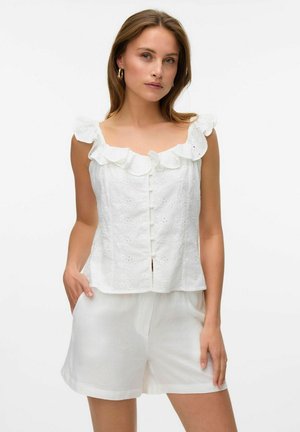 Top sans manches blanc en tissu à œillets, avec un col à volants et une fermeture à boutons à l'avant. Associé à un short blanc ample.