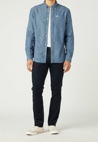 Denim button-up shirt in light blue, with a chest pocket and white buttons; paired with dark pants and light sneakers. Casual fit.