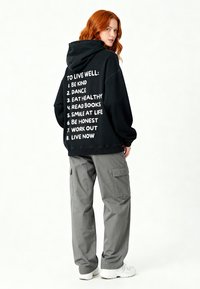 Felpa nera con testo bianco sul retro che elenca otto pratiche per il benessere. Abbinata a pantaloni cargo grigi e sneakers bianche.