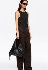 Czarna skórzana torebka z frędzlami i ćwiekami, zestawiona z ciemnym topem na ramiączkach i szerokimi brązowymi spodniami. Matowe faktury i elegancki design.