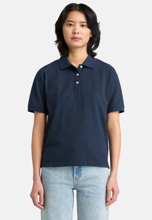 Junge Person mit schulterlangem schwarzen Haar, die ein marineblaues Poloshirt mit kurzen Ärmeln und hellblaue Jeans trägt und vor einem einfarbigen Hintergrund steht.