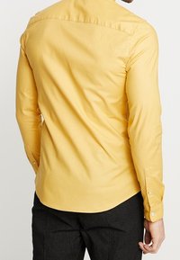 Mężczyzna w dopasowanej, żółtej koszuli z długim rękawem i czarnych spodniach, pokazany z tyłu na tle jednolitym.