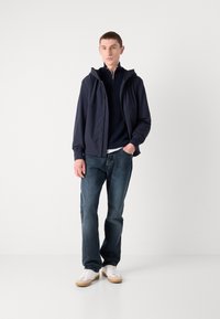 Marine windbreaker met rits aan de voorkant en capuchon, gecombineerd met een donkerblauwe gebreide trui, denim jeans en witte sneakers met beige accenten.