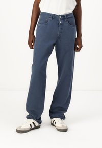 Blauwe denim jeans met een losse pasvorm, vijf zakken, twee knopen aan de voorkant en zichtbare stiksels. Gecombineerd met grijze en witte sneakers.