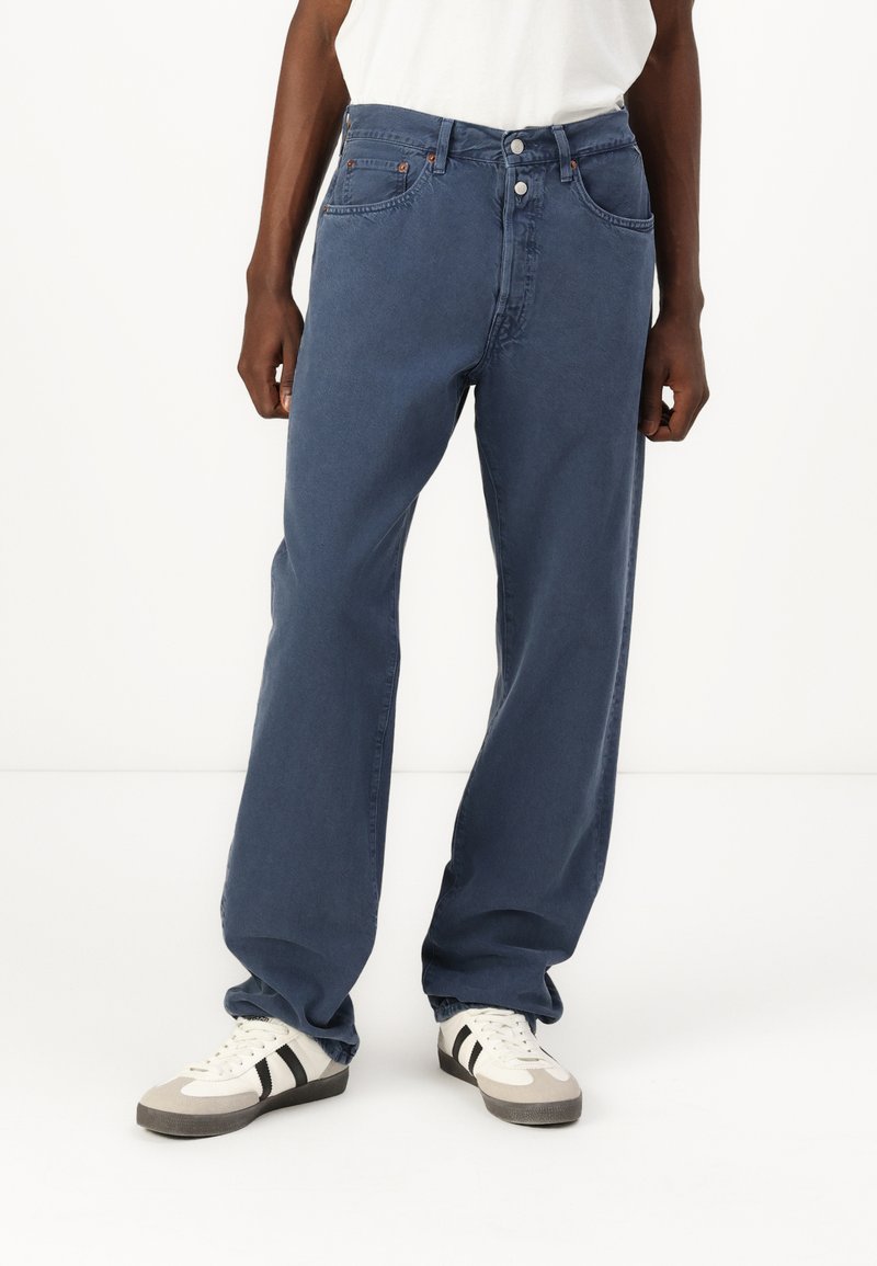 Blauwe denim jeans met een losse pasvorm, vijf zakken, twee knopen aan de voorkant en zichtbare stiksels. Gecombineerd met grijze en witte sneakers.