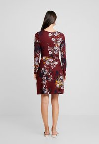 Mörkröd långärmad klänning med ett blommönster i vitt, gult och marinblått. Åtsittande midja och utsvängd kjol, tillverkad av mjukt, strukturerat tyg.