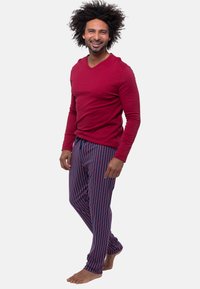 Röd långärmad t-skjorta med v-ringning som är matchad med blå pyjamasbyxor med vertikala röda och vita ränder. Modellen står barfota och ler.