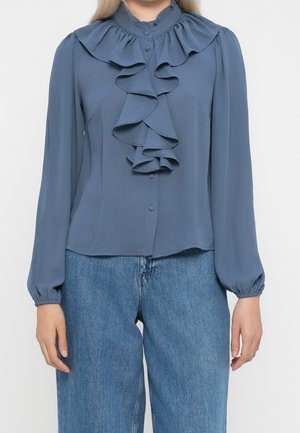 Femme portant un chemisier bleu poussiéreux à manches longues avec un devant volanté et un col, associé à un jean bleu taille haute.