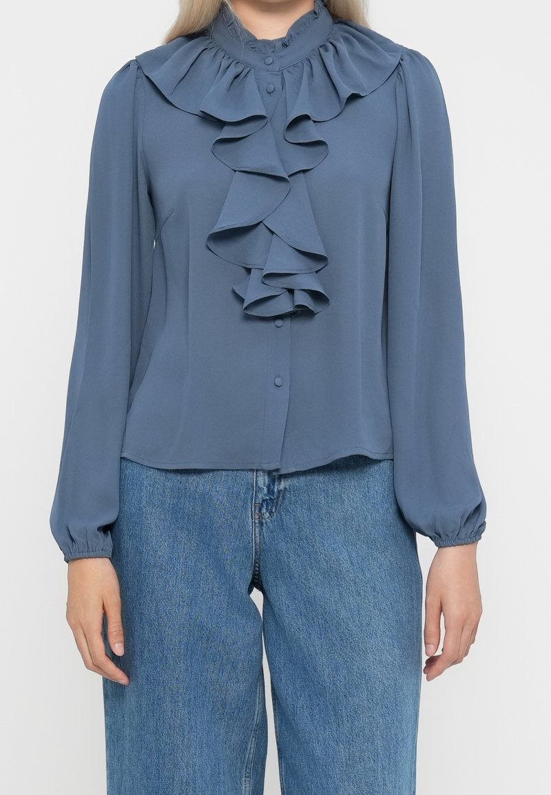 Femme portant un chemisier bleu poussiéreux à manches longues avec un devant volanté et un col, associé à un jean bleu taille haute.