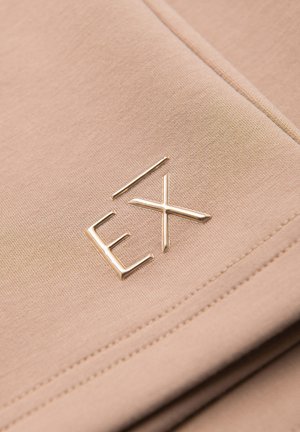 Gros plan sur un tissu beige à texture lisse avec des lettres métalliques dorées en relief « EX » cousues sur la surface.