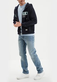 Felpa blu marina con zip e testo frontale, abbinata a una maglietta azzurra chiara e jeans in denim chiaro. Sneakers bianche completano il look casual.