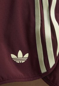 Rødbrunt stof med små perforeringer, med tre lodrette off-white striber og et off-white Adidas Trefoil-logo.