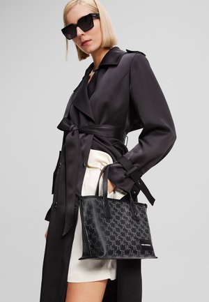 Borsa a mano in pelle nera con un motivo testurizzato di piccole forme geometriche, design rettangolare e doppi manici. Indossata con un trench coat scuro.