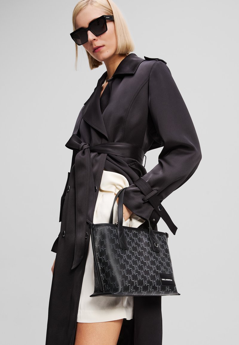 Svart läderhandväska med ett texturerat mönster av små geometriska former, rektangulär design och dubbla handtag. Bärs med en mörk trenchcoat.