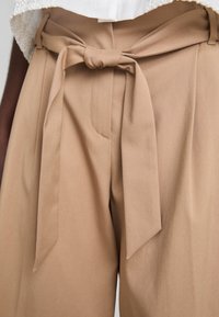 Beige high-waisted trousers with a tied belt detail, pleats at the front, and a smooth fabric texture. Features side pockets and minimal stitching.