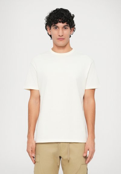 Joven con cabello oscuro y rizado, vistiendo una camiseta blanca de manga corta y pantalones cargo beige, de pie contra un fondo blanco.