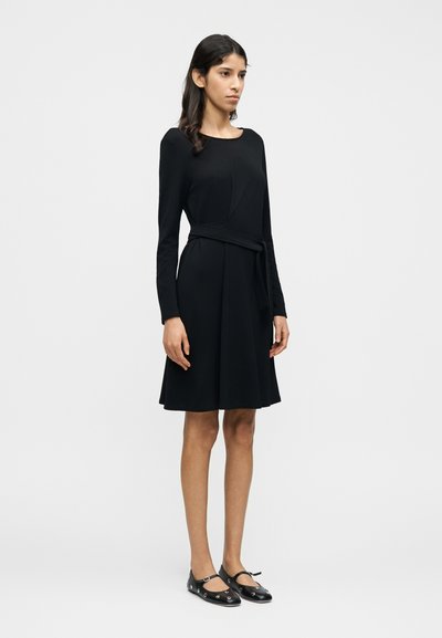 Μαύρο φόρεμα με μακριά μανίκια, εφαρμοστό μπούστο και φουσ κωτή φούστα, συνοδευόμενο από μαύρα επίπεδα παπούτσια με λουράκια στον αστράγαλο και διακοσμητικές λεπτομέρειες.