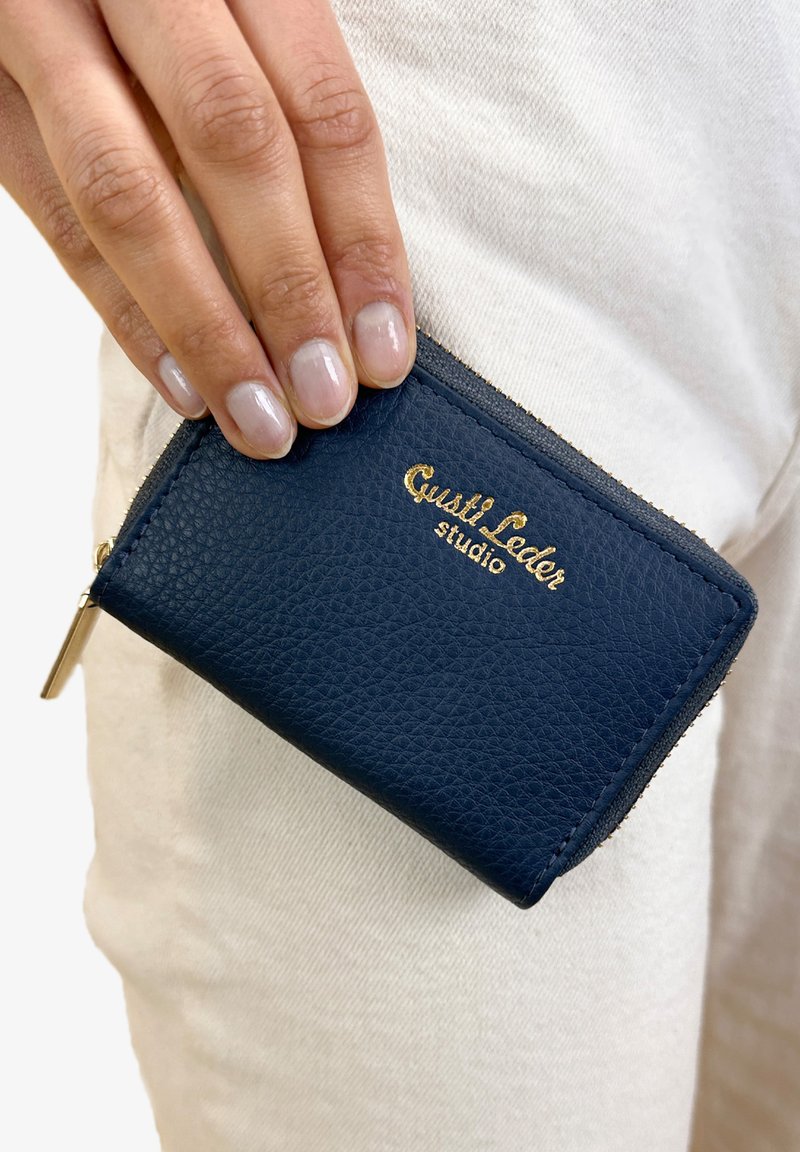 Navy blue leather card holder with a textured finish, gold embossed logo, and a zipper. Held in a hand against a light background.