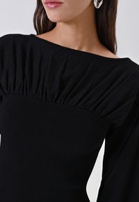 Donna che indossa un top fitted nero a maniche lunghe con scollatura a spalle scoperte e dettagli di tessuto arricciato sul petto, insieme a orecchini a cerchio argentati.