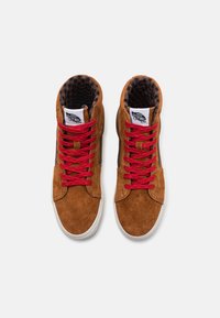 Zapatillas altas de ante marrón con cordones rojos estampados, suelas de goma blanca y un forro a cuadros en negro y marrón. Presenta un parche de logo en la lengüeta.