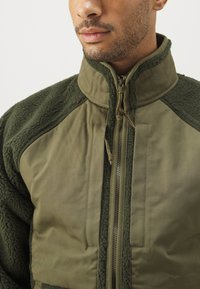 Jachetă verde măslin care combină fleece-ul moale și texturat cu un material neted, având guler înalt și fermoar frontal.