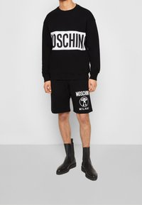 Felpe nere e pantaloni corti con branding bianco "MOSCHINO", abbinati a stivali neri fino alla caviglia. Il materiale sembra morbido con una vestibilità casual.
