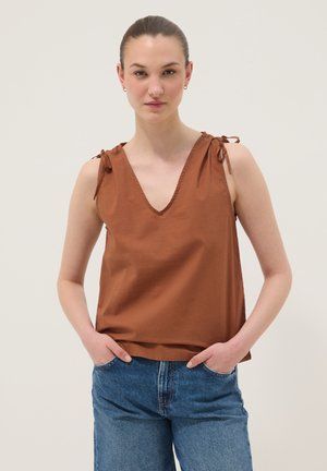 Jeune femme portant un haut marron sans manches avec épaules nouées et un jean bleu, debout les mains dans les poches devant un fond uni.