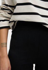 Camisa de manga larga a rayas en crema y negro, combinada con pantalones negros de cintura alta. El antebrazo presenta un tatuaje a rayas en verde y azul.