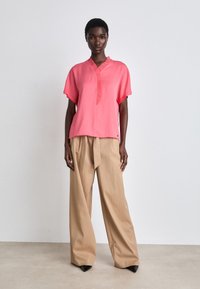 Rosa kurzärmlige Bluse mit V-Ausschnitt und Faltenoptik, kombiniert mit hellbeigen weit geschnittenen Hosen und spitzen schwarzen High Heels.