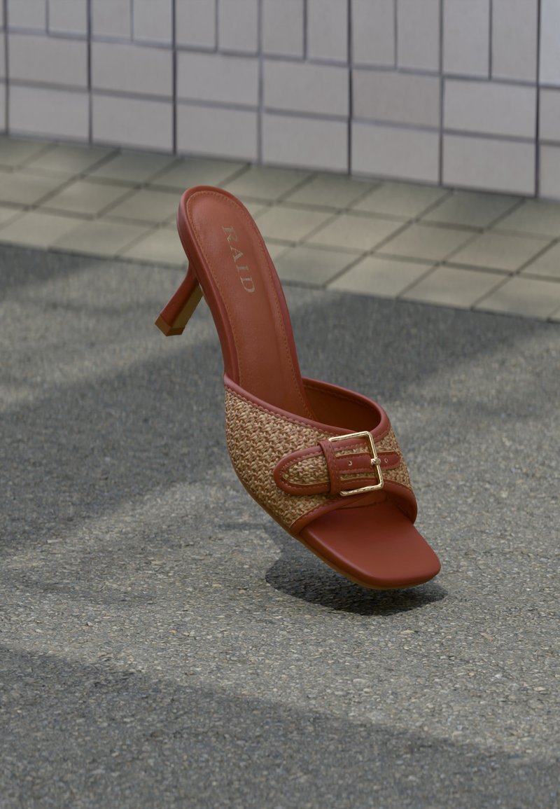 Mule à talon haut marron avec une lanière tissée et une boucle dorée, reposant sur une surface pavée extérieure près d'un mur carrelé en arrière-plan.