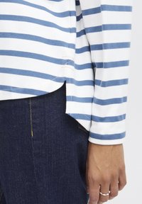 Striped long-sleeve shirt in white and blue cotton, featuring a side slit and a curved hem, worn with dark denim jeans.