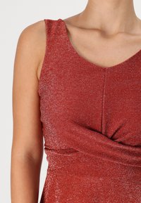Robe rouge sans manches avec décolleté en V, présentant une taille plissée et une texture pailletée. Tissu lisse avec brillance et détails subtils.