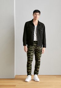 Jachetă din denim negru peste un tricou alb, asortată cu pantaloni verde camuflaj și adidași albi. Design simplu, formă ajustată și contrast textural.