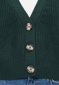 Zielony sweter w robionym na drutach fasonie z dekoltem w serek, ozdobiony trzema okrągłymi guzikami w jasnych i ciemnych odcieniach, połączony z niebieskim dżinsem na dole.